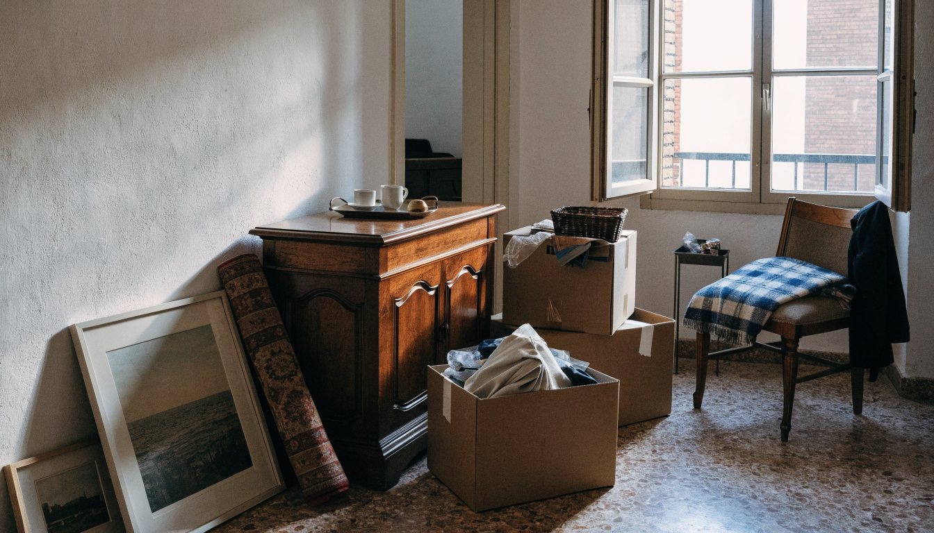 Stanza abitata prima dello sgombero: una credenza in legno, scatoloni mezzi pieni, quadri e un tappeto arrotolato appoggiati al muro