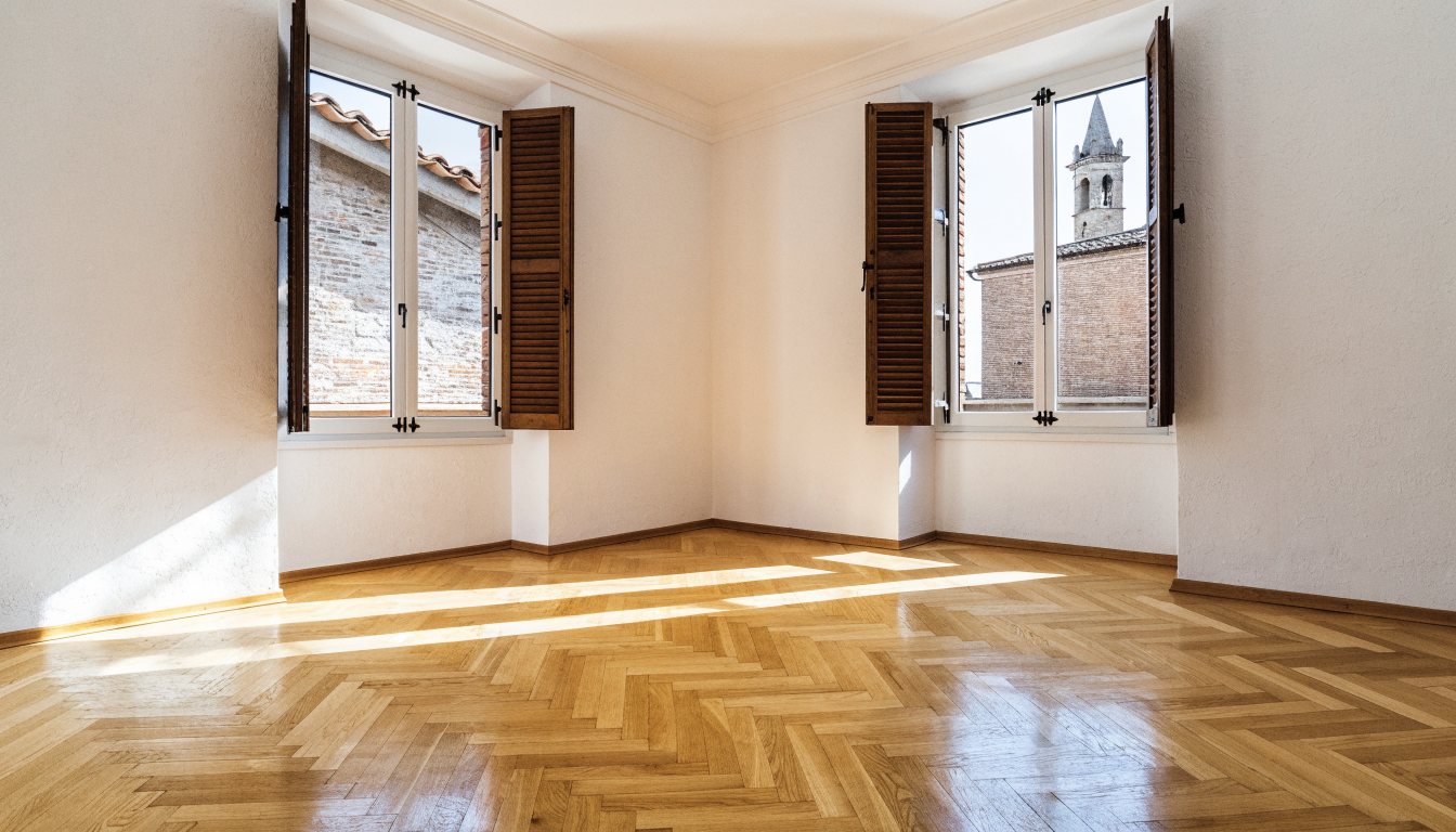 Salone vuoto e luminoso con parquet a spina di pesce e due finestre aperte sui tetti e su un campanile